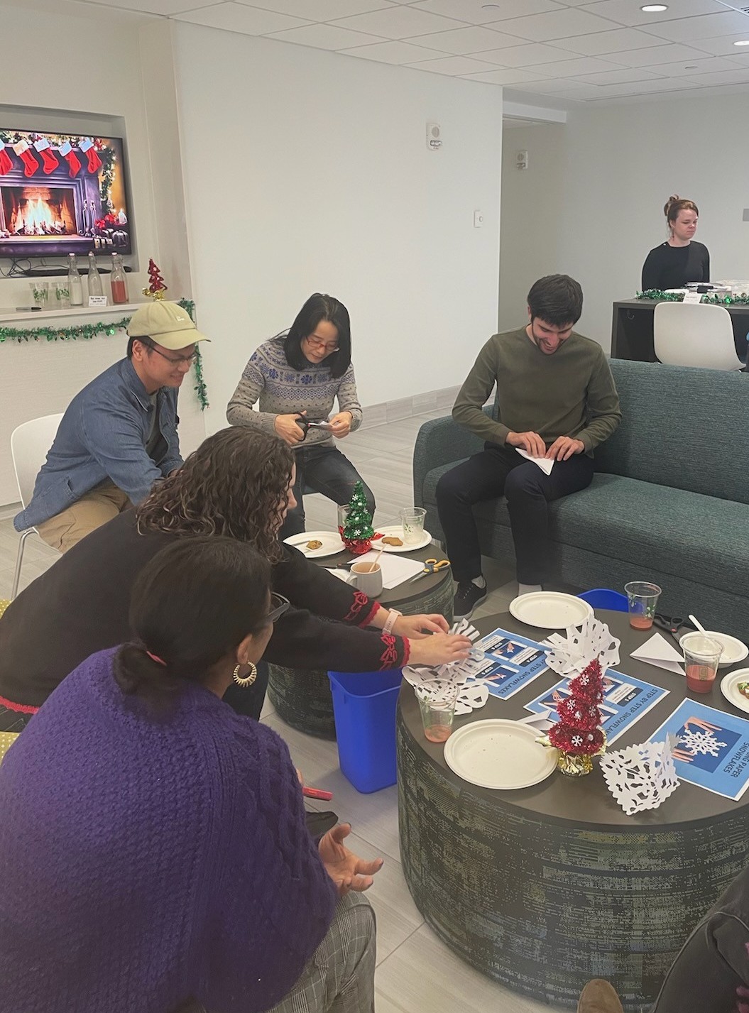 Five people sit around a table making paper snowflake crafts, one person stands further away