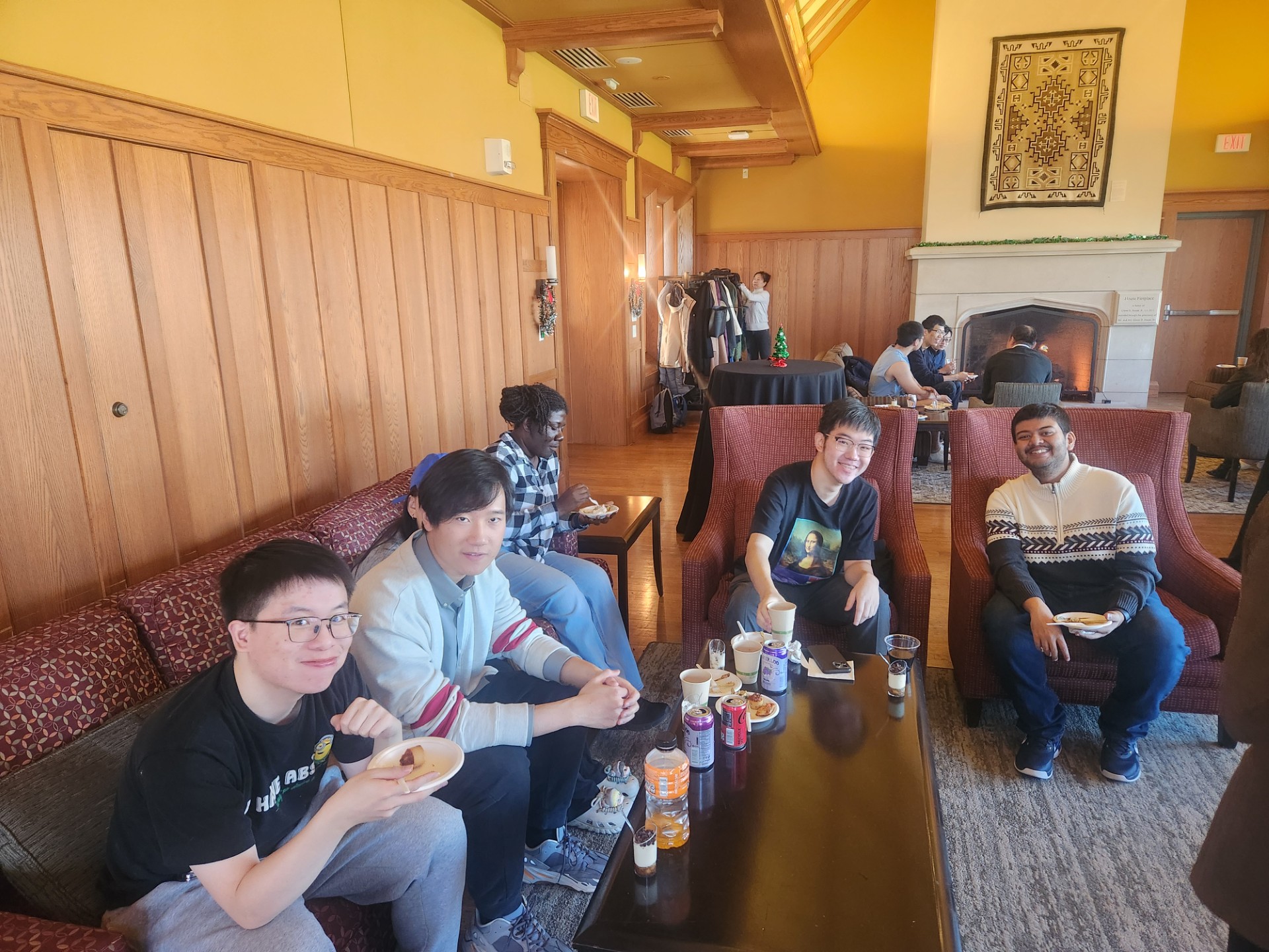 Five people sitting on couches smile at the camera, in the background there are more people