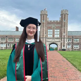 Qingwen (Vicky) Zeng stands in front of Brookings Hall in her graduation robes